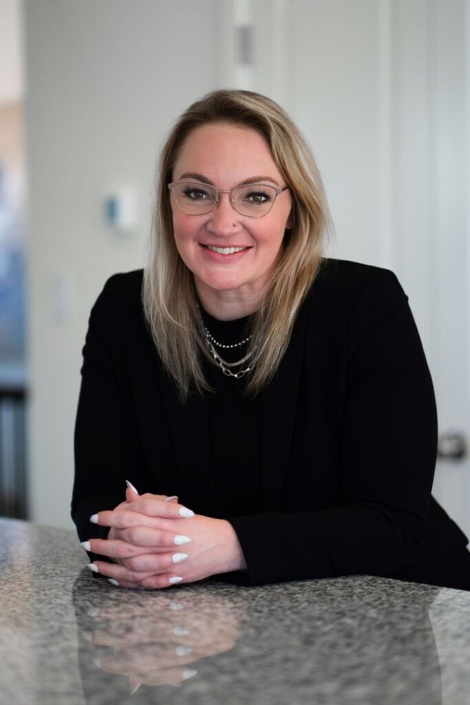 A women with blonde hair wearing a black blouse and glasses on a white background.