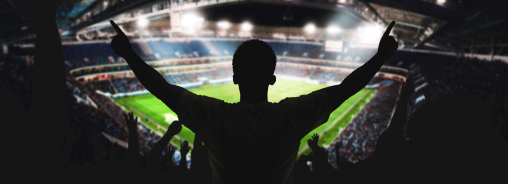 A silhouetted figure with arms stretched up and out to their side, viewed from the back. They appear to be standing on the top section of a sports stadium, which stretches out in front of them.